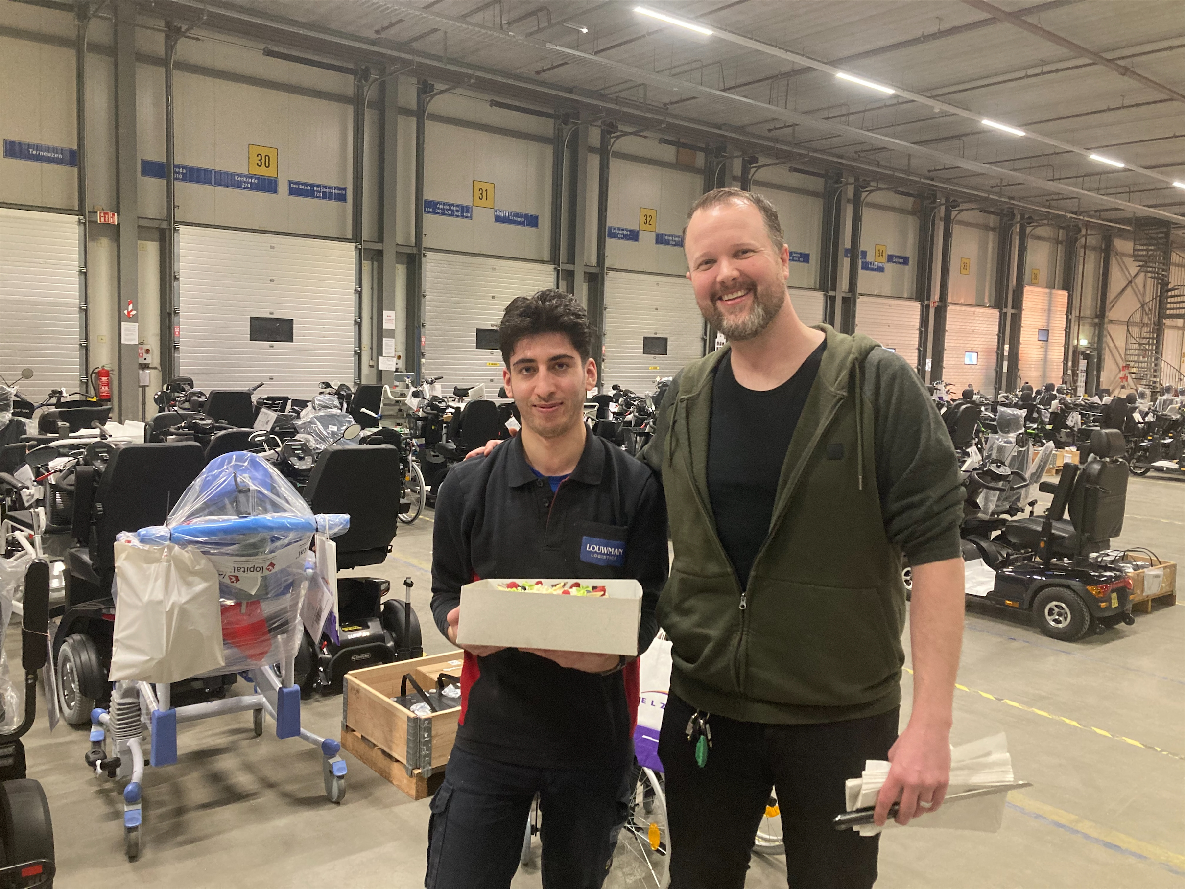 Murat en zijn werkgever staan in het magazijn bij elkaar, omringt voor scootmobiels. Murat heeft een taart vast. 