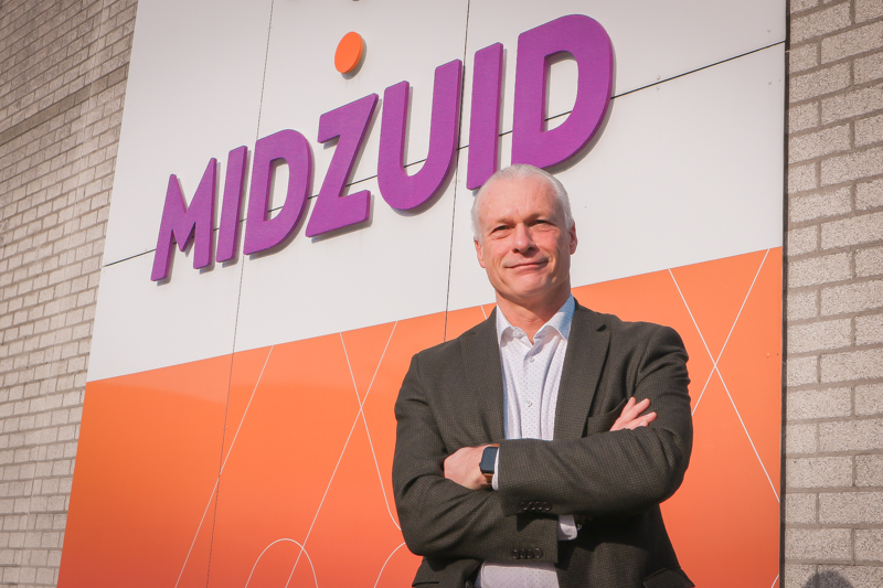Peter kijkt lachend in de camera en staat met zijn armen over elkaar voor de muur bij de ingang van het pand waar groot het logo van MidZuid op staat.